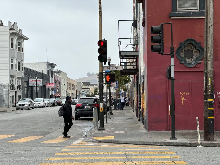 A person crosses a city street at a crosswalk with a red traffic light and a "16th St" sign visible on a cloudy day.