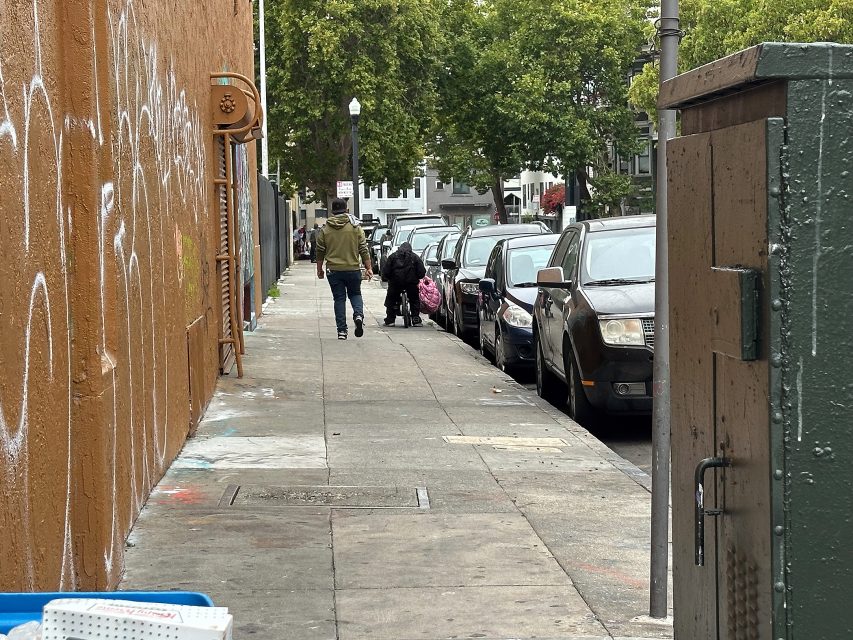 A person walks down a city sidewalk lined with parked cars and graffiti, while another person with bags stands further ahead. Trees and buildings are visible in the background.