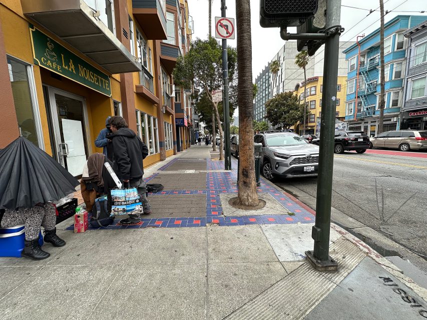 People gather with belongings on a city sidewalk near a café, with parked cars and buildings lining the street on an overcast day.