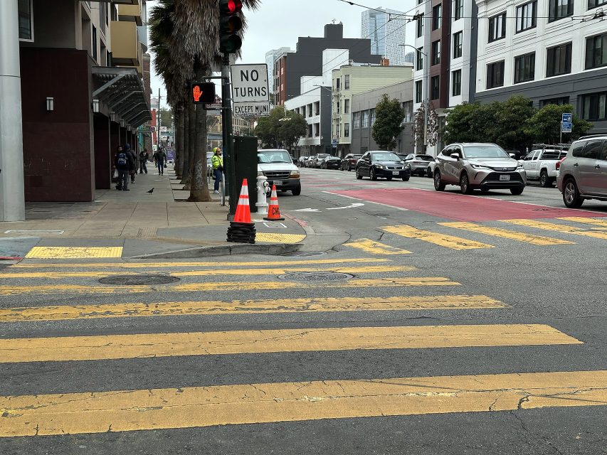 A crosswalk with yellow stripes, traffic cones on the corner, and a "NO TURNS" sign. Several cars and buildings are visible in the background.