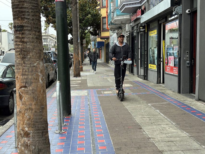 A man rides an electric scooter on a city sidewalk lined with shops, parked cars, pedestrians, and trees.