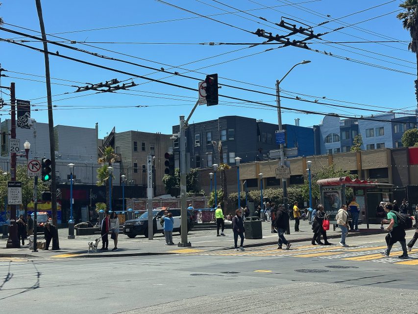Intersection with pedestrians crossing, traffic signals, power lines overhead, and buildings in the background on a sunny day.