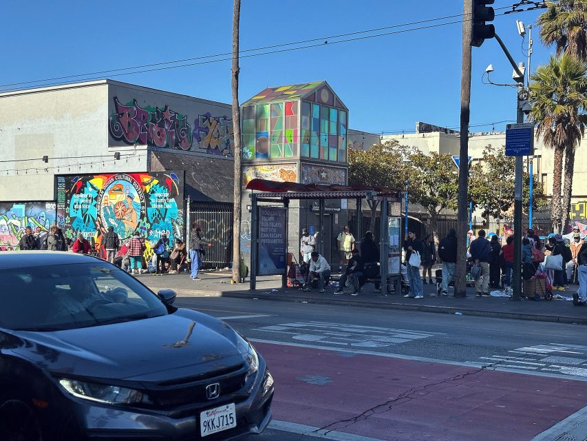 People gather near a bus stop on a city street lined with graffiti-covered buildings and a colorful structure under a clear blue sky.