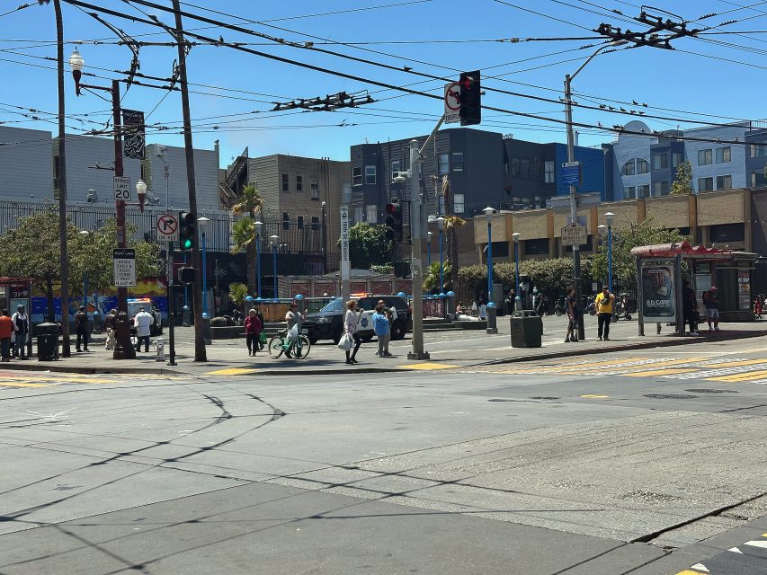 A busy urban intersection with pedestrians, cyclists, and people waiting at a bus stop; overhead wires and buildings are visible under a clear sky.