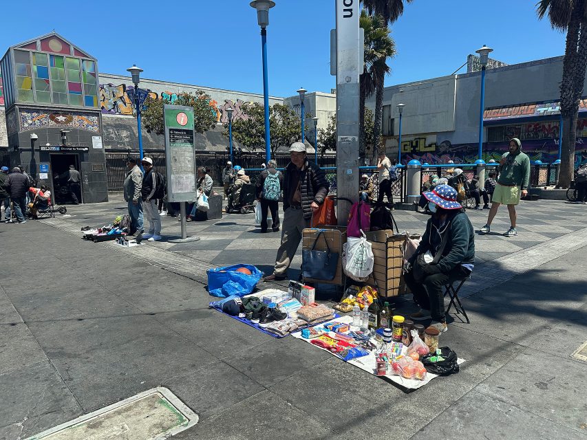 Two street vendors sell various goods laid out on the sidewalk in a busy urban plaza, with people and graffiti-covered buildings in the background.