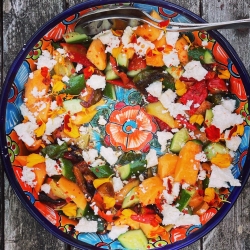 A colorful salad with diced vegetables, cheese crumbles, and edible flowers in a vibrant, patterned bowl on a rustic wooden surface.