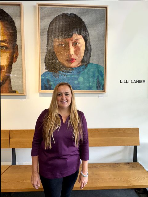 A woman with long blonde hair stands in front of a wooden bench and a wall displaying pixelated portraits, including one with the name "Lilli Lanier" beside it.