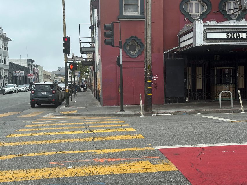 A crosswalk with yellow stripes leads to a red building on a cloudy day; traffic lights are red, and cars are parked along the street.
