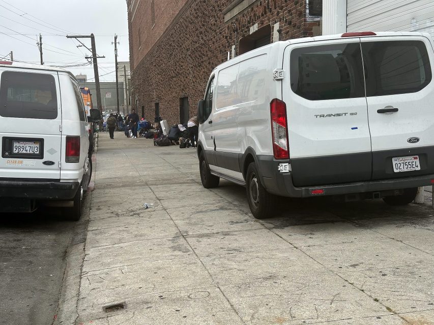 Two white vans are parked on a city street while a group of people with belongings gather near a brick building in the background.
