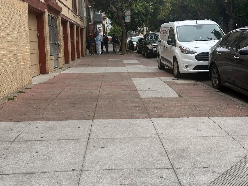 A group of people stand on a sidewalk near parked cars and buildings on an overcast day in an urban neighborhood.