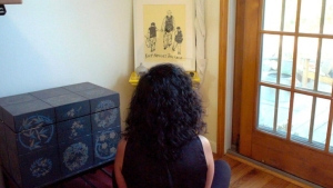 A person with curly dark hair sits on the floor facing a framed picture on a shelf near a glass door and a blue decorative chest.