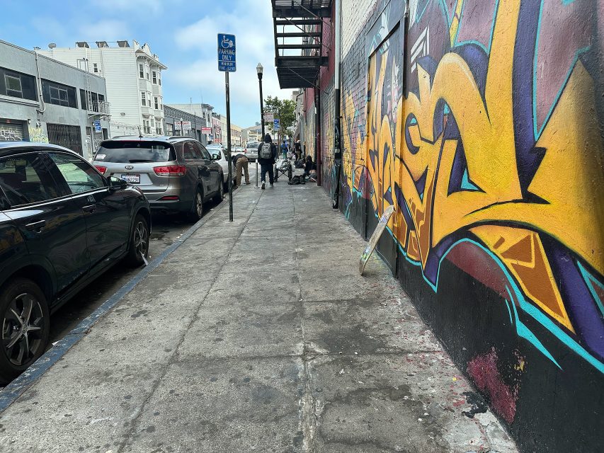 Urban sidewalk with parked cars on the left, colorful graffiti on the right wall, and several people walking or standing in the distance under cloudy skies.