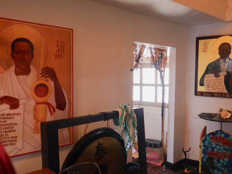 A room with two religious-style paintings of Black men on the walls, a large gong in the foreground, and light streaming through a window in the background.
