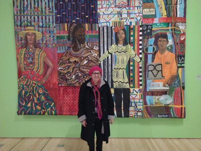 A person in a red hat and scarf stands in front of a colorful textile artwork depicting four figures wearing patterned clothing.