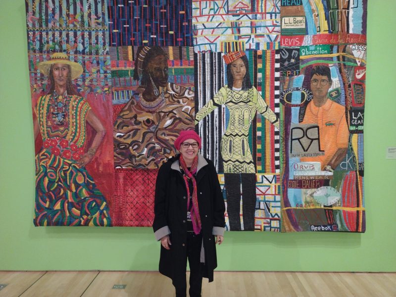 A person in a red hat and scarf stands in front of a colorful textile artwork depicting four figures wearing patterned clothing.