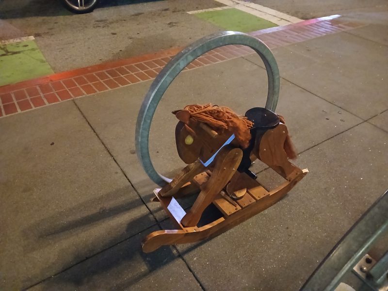 A wooden rocking horse is locked to a metal bike rack on a sidewalk at night.
