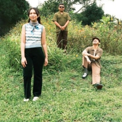 Three people pose outdoors on a grassy area with plants and trees in the background; one stands in front, one sits on the grass, and one stands further back.