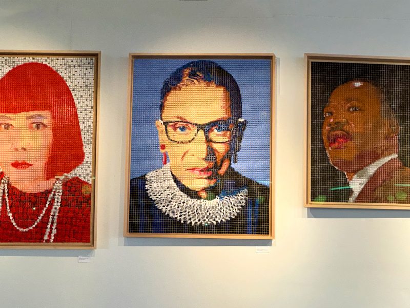 Three framed portraits made from small colored tiles hang on a white wall, depicting individuals with distinct clothing and accessories.