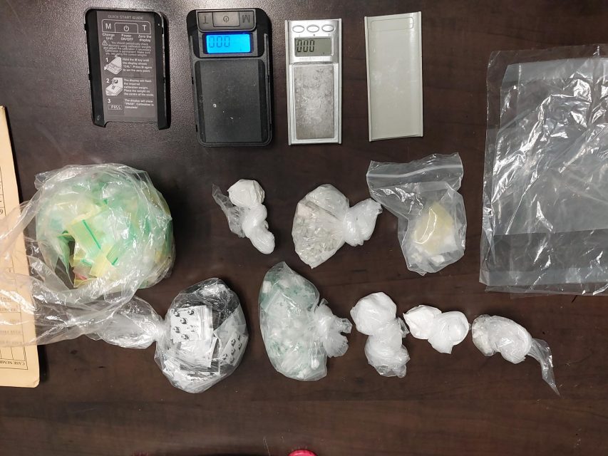 Several digital scales and multiple plastic bags containing white crystalline substances are arranged on a dark surface, along with empty plastic bags and a brown envelope.