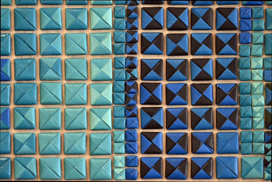 A grid of origami squares and triangles in various shades of blue and turquoise is arranged in an orderly pattern.