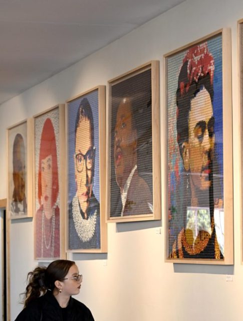 A woman wearing glasses looks at framed mosaic portraits of famous figures displayed on a white wall in an art gallery.