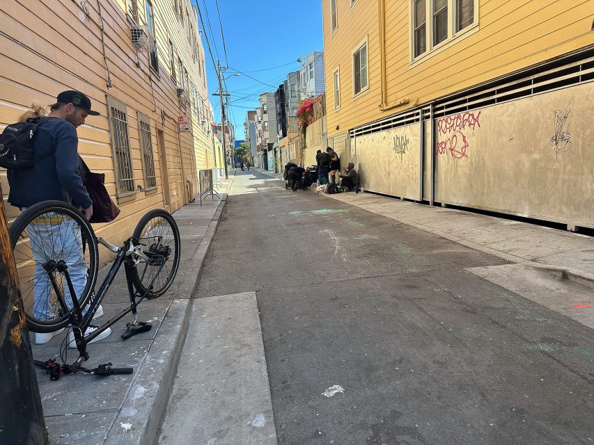 A person repairs a bicycle on the left side of a narrow urban alley, while a group of people gathers near a building further down the alley.