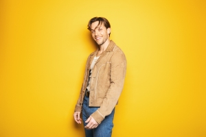 A man wearing a tan suede jacket, white shirt, and blue jeans stands smiling against a solid yellow background.