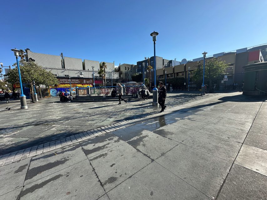 A city plaza with a few people walking and sitting, surrounded by buildings, trees, lamp posts, and murals under a clear blue sky.
