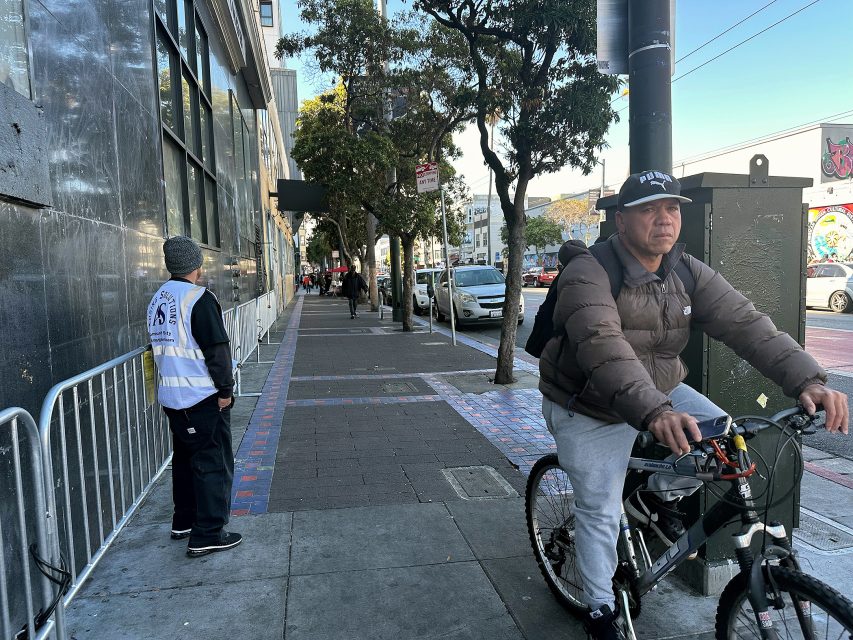 A man rides a bicycle on a city sidewalk while another man in a reflective vest stands near a building. Cars are parked along the street and pedestrians walk in the distance.