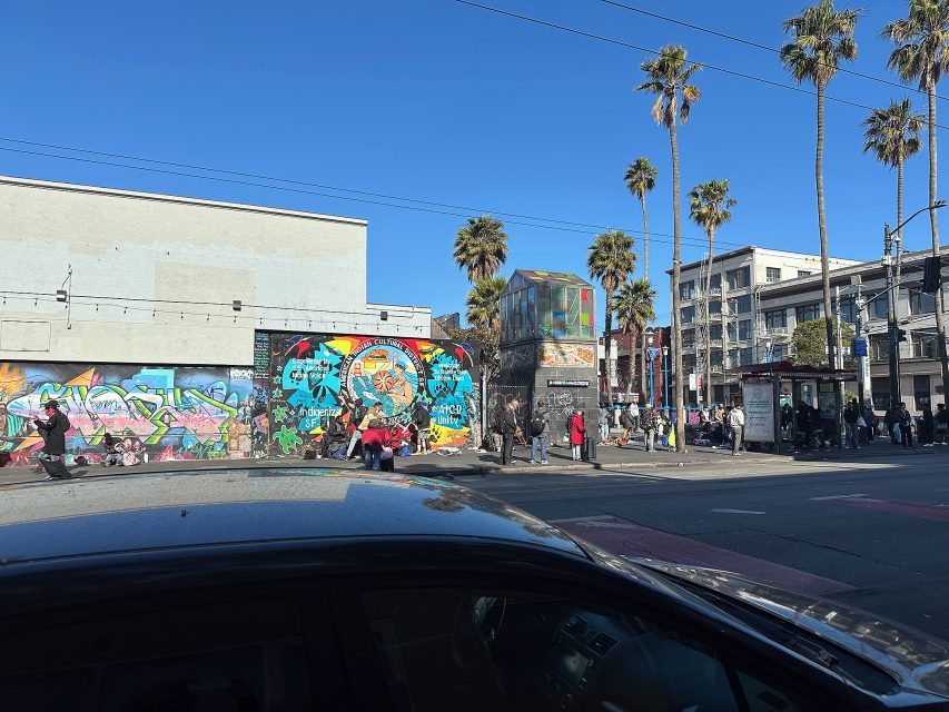 People gather near colorful graffiti murals and palm trees on a sunny day at an urban street corner, with cars and buildings in the background.