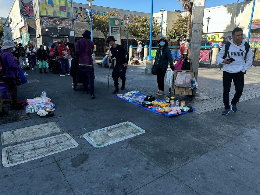 People gather around street vendors displaying various goods on the ground in an urban outdoor market setting with colorful murals in the background.
