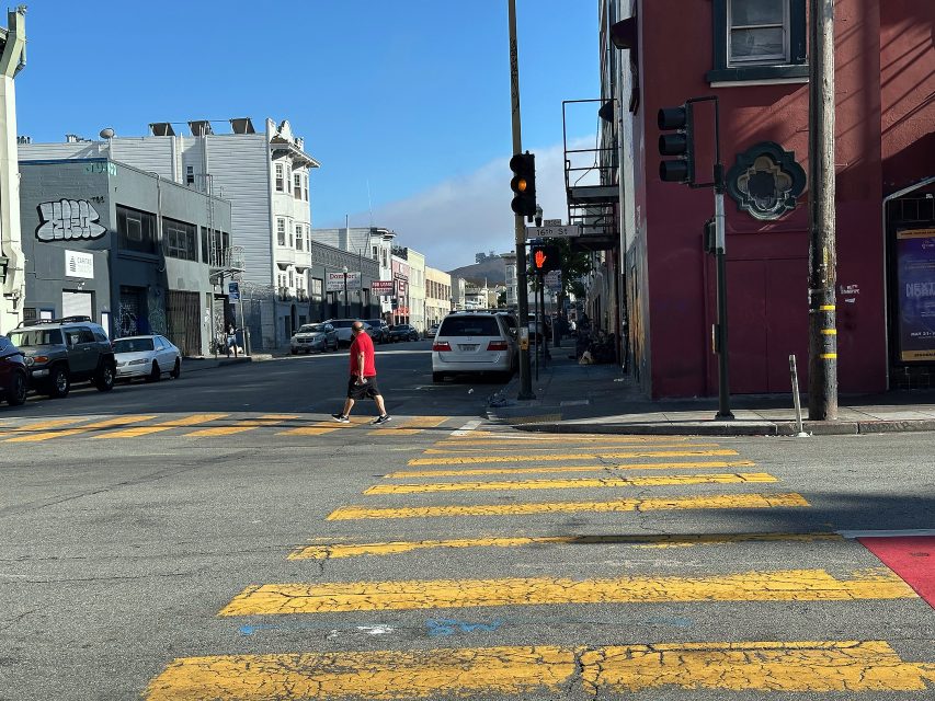 A person in a red shirt crosses a street at a crosswalk in an urban area with buildings, cars, and a traffic light visible.