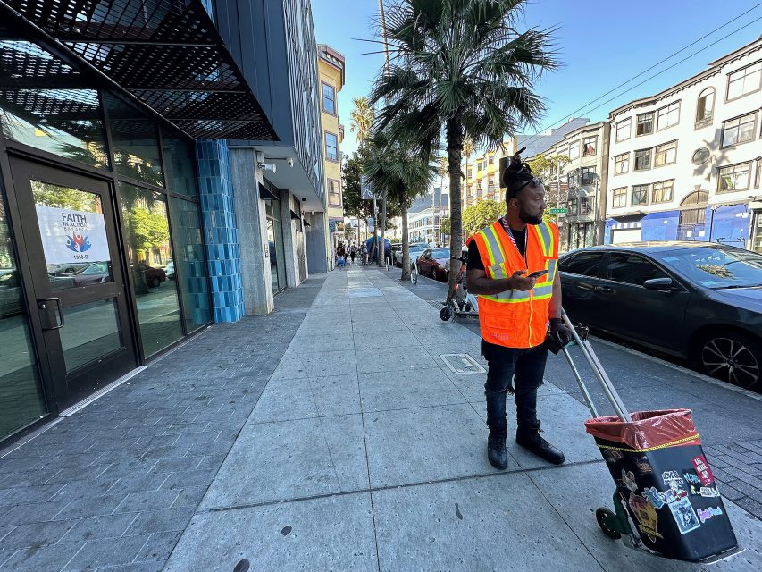 A man in a fluorescent safety vest stands on a city sidewalk, holding a phone and pushing a wheeled trash bin. Cars and buildings line the street.