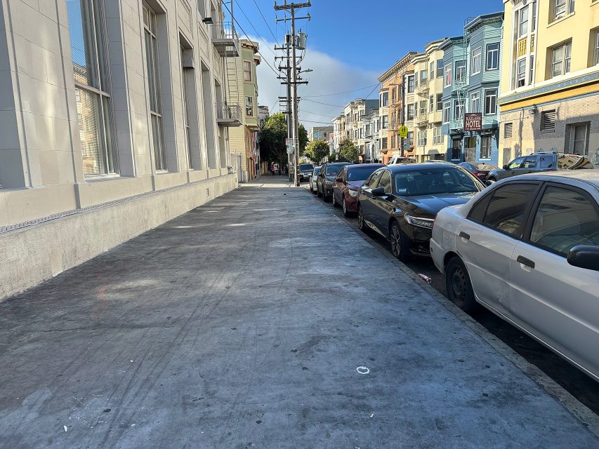 A city sidewalk lined with parked cars and multi-story residential buildings on a clear day.