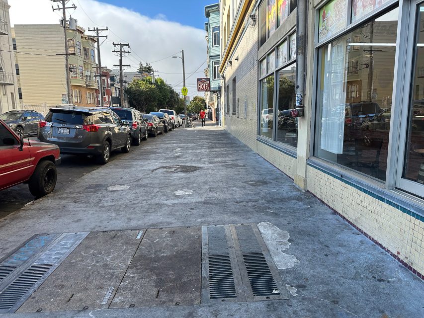 A city sidewalk lined with parked cars on the left and storefronts on the right, with a few pedestrians visible in the distance under a partly cloudy sky.
