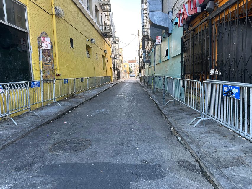 A narrow urban alley with metal barricades lining both sides, yellow and blue buildings, and no people visible.