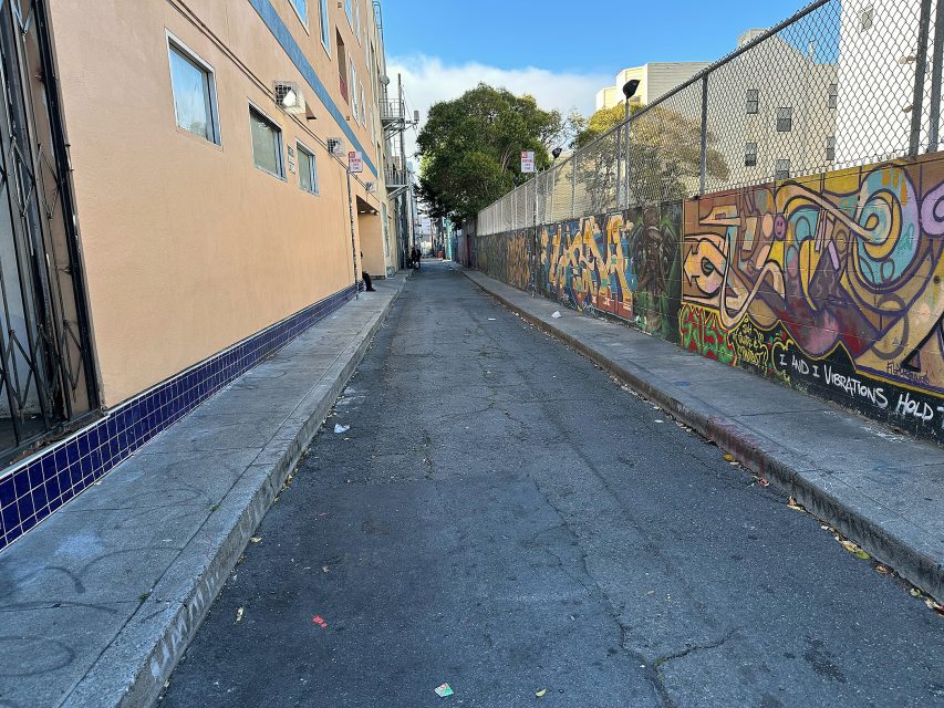 A narrow urban alleyway with graffiti on the right wall, a beige building on the left, and litter scattered along the pavement.