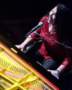 A person in a red top sits at a grand piano, singing into a microphone, with piano strings and keys visible from above.
