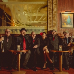 Six people dressed in formal attire sit at two small round tables with drinks, posing indoors against a decorated wall with framed art and a large frosted glass panel.
