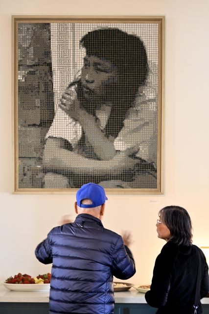 Two people stand in front of a table with food, conversing beneath a large monochrome portrait of a woman displayed on the wall.