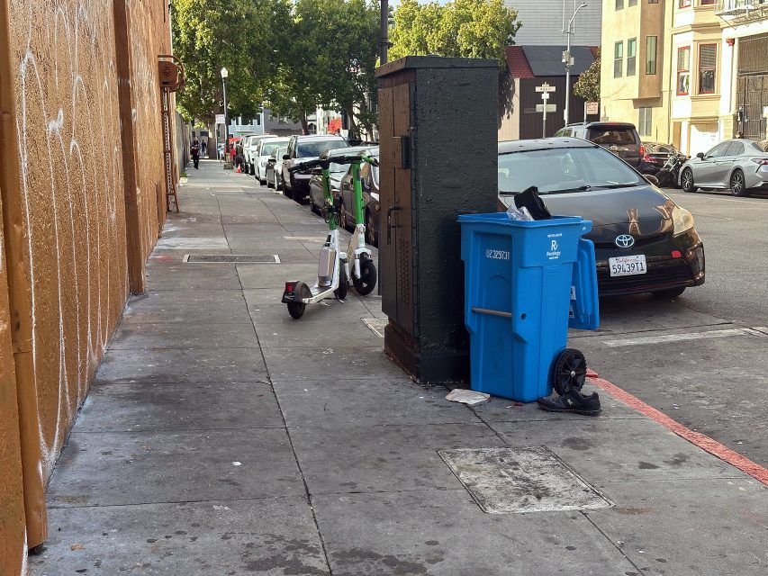 A city sidewalk with a recycling bin, black trash bin, and electric scooter near the curb; parked cars and buildings line the street.