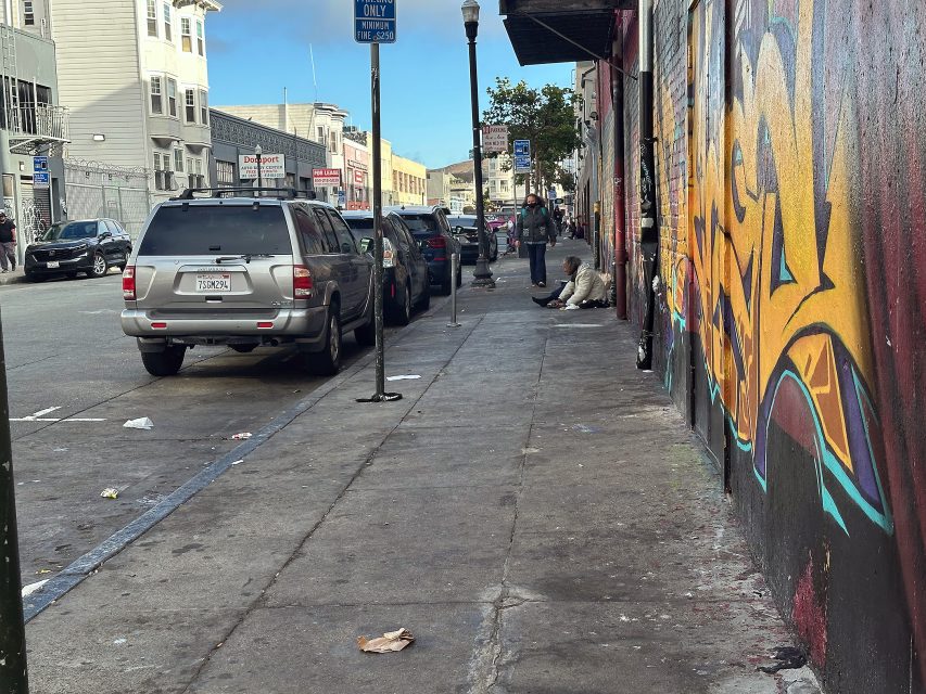 A city sidewalk with parked cars, a handicap parking sign, colorful graffiti on the wall, and a person sitting on the ground near a building.