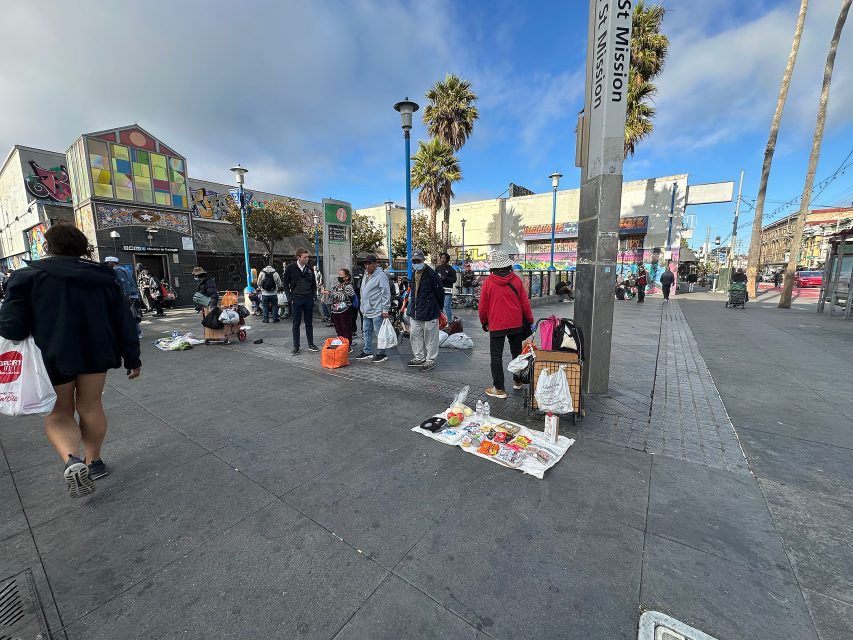 People are gathered on a city sidewalk near street vendors selling various items laid out on blankets, with colorful buildings and palm trees in the background.