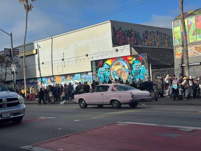 A light purple classic car drives past a crowd of people standing by a graffiti-covered building on a city street.