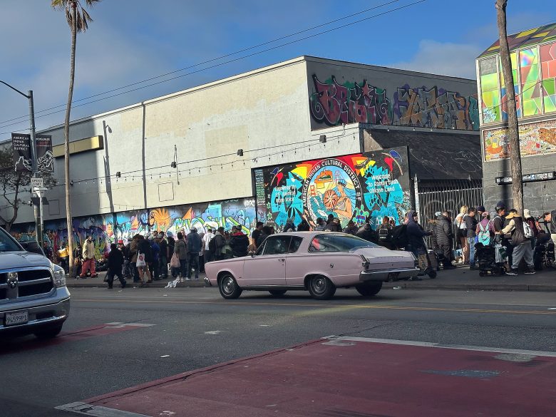 A light purple classic car drives past a crowd of people standing by a graffiti-covered building on a city street.