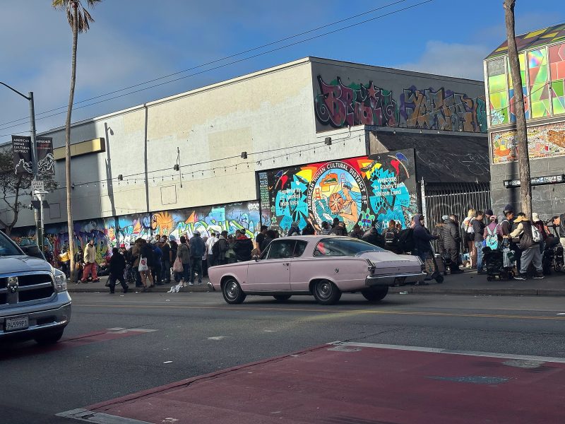 A light purple classic car drives past a crowd of people standing by a graffiti-covered building on a city street.
