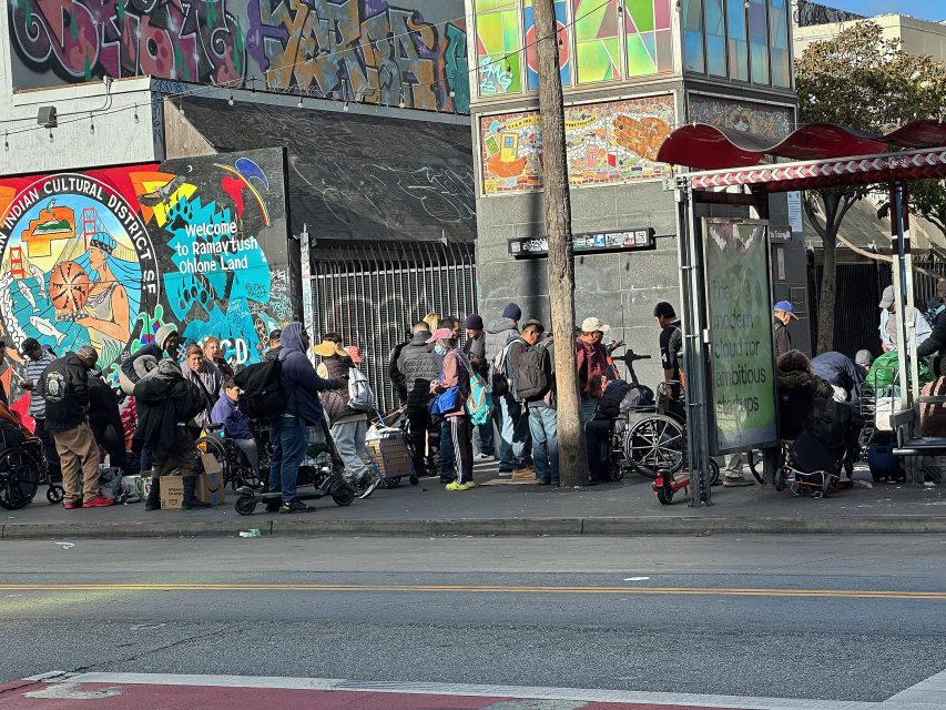 A group of people stand in line with carts and bags near a colorful mural and a bus stop on a city sidewalk.