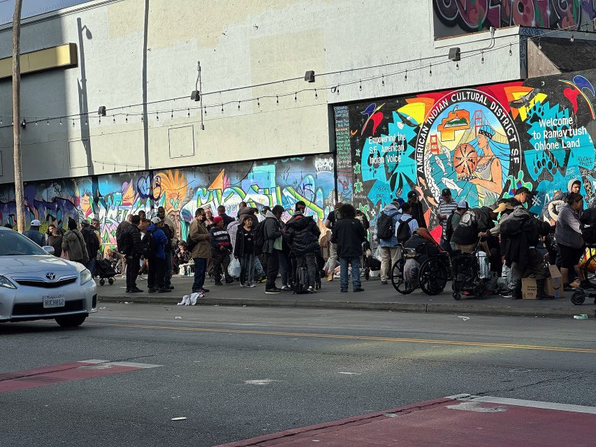 A large group of people gathers on a sidewalk in front of a colorful mural in an urban area; some stand, others use wheelchairs or carry bags.