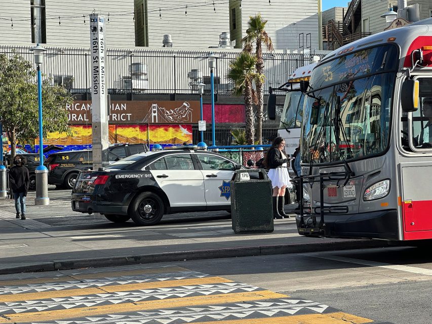 A city street scene with a police car, a bus, a person standing on the sidewalk, and a mural reading "American Indian Cultural District" in the background.