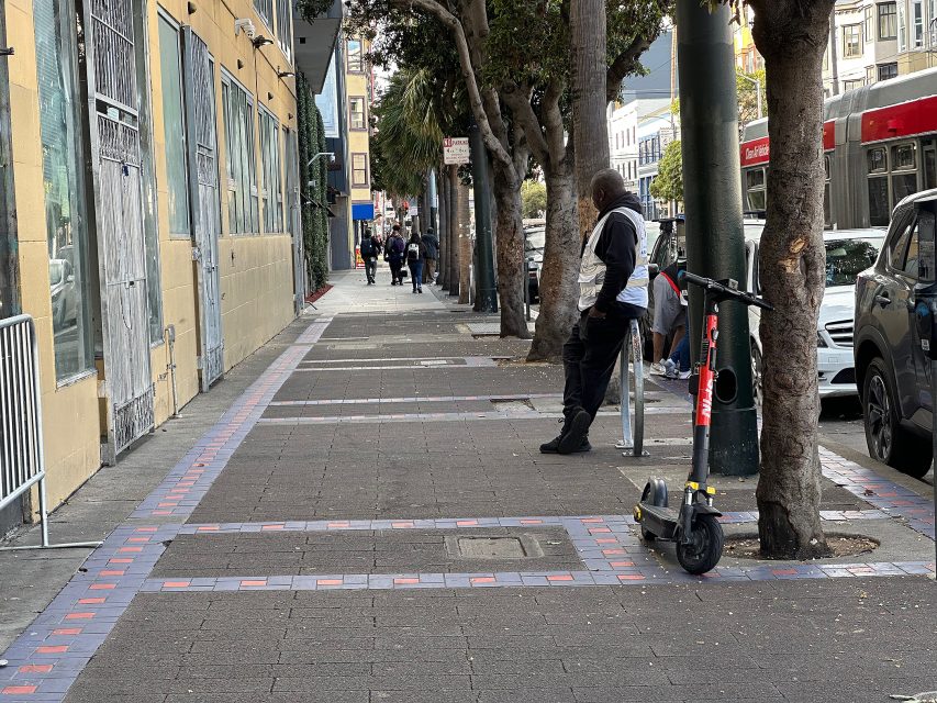 A man leans against a tree on a city sidewalk next to an electric scooter; pedestrians and vehicles are visible in the background.